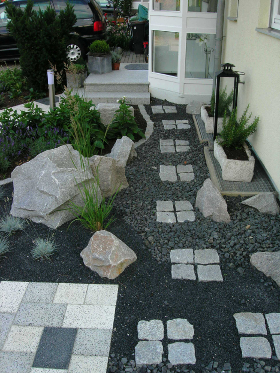 Schwarzer Basaltschotter und graue, formwilde Granitsteine führen mit Trittplatten zum Eingang